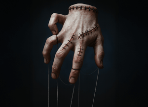 Una mano umana cucita con grosse suture, sospesa come una marionetta da fili sottili, su sfondo scuro. L’immagine evoca un’atmosfera inquietante e surreale.